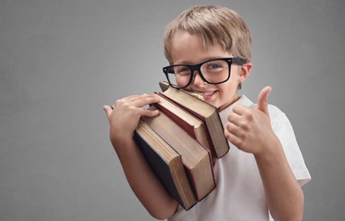 Ein kleiner Junge hält einen Stapel Bücher auf dem Arm, lächelt in die Kamera und zeigt einen Daumen nach oben.